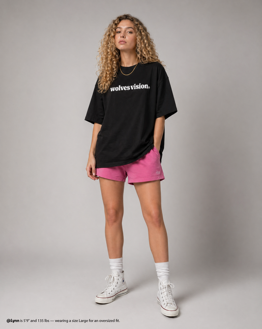 Person wearing a black oversized t-shirt with 'wolves vision' text, pink shorts, and white sneakers on a gray background.