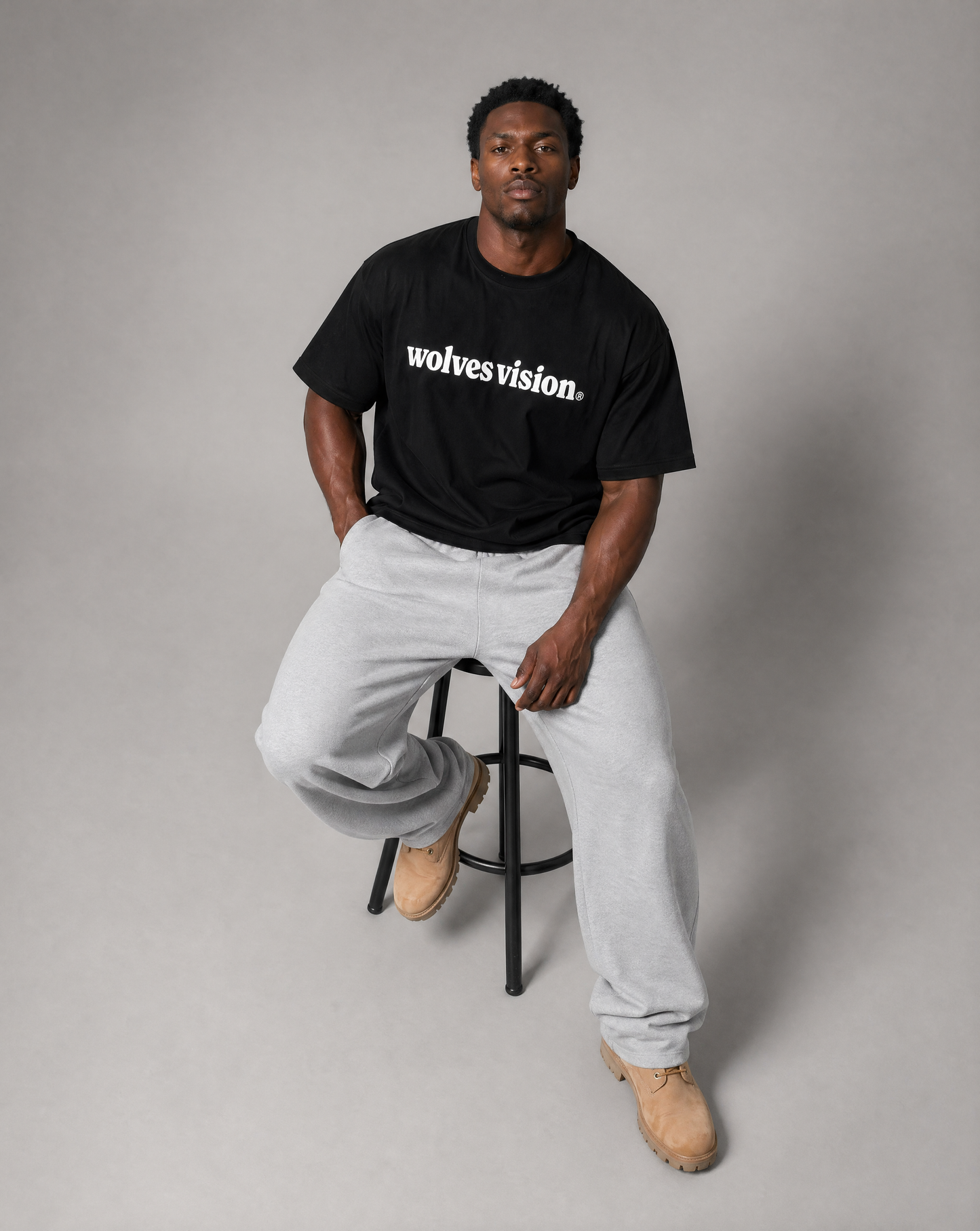 Black man on stool sitting uo with one foot on floor and other foot on the stool bar looking up at camera with black shirt on saying "wolves vision"