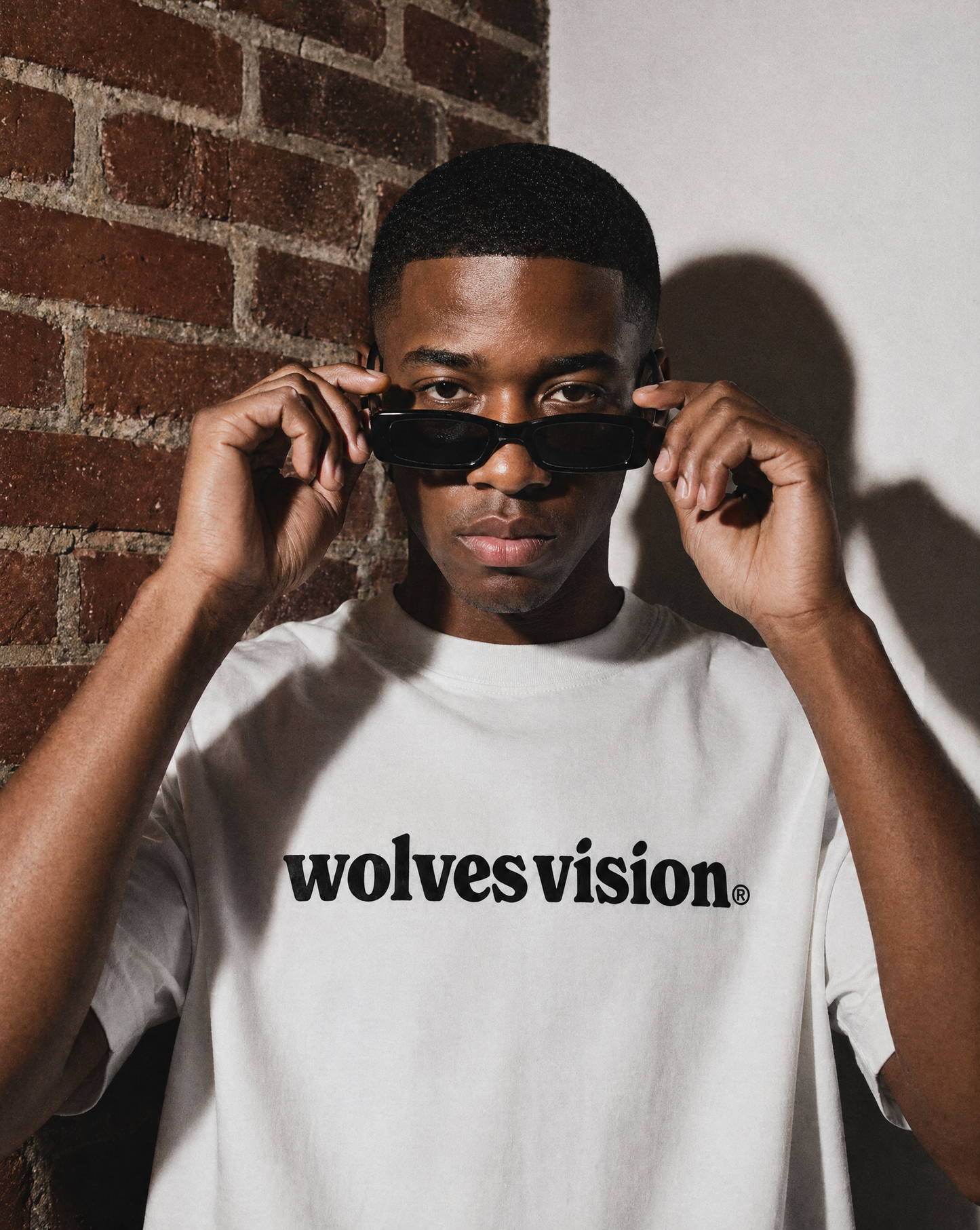 Person wearing a white t-shirt with 'wolvesvision' text, holding sunglasses, against a brick wall.